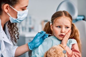 Child in dental chair holding teddy bear, showing concern during pediatric tooth extraction procedure with dentist in mask and gloves.