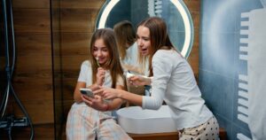 Two girls in a bathroom, one showing the other something on a smartphone, emphasizing the importance of engaging with children about oral hygiene habits.