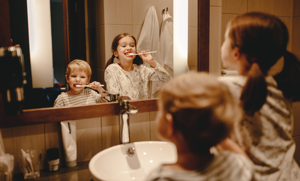 Children brushing their teeth together in a bathroom mirror, promoting healthy dental habits and parental involvement in oral hygiene.